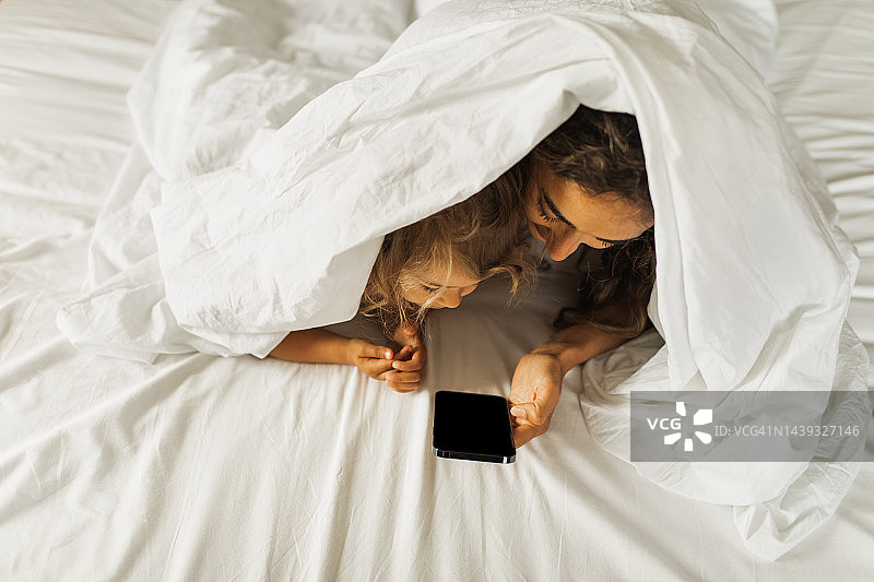 快乐的母女俩躲在床上的毯子下，用手机看动画片图片素材