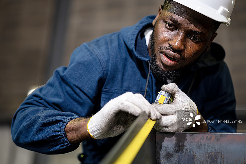 施工梁结构与结构跟踪精度。一个非裔美国金属工人在工厂用不锈钢卷尺测量建筑梁的低角度视图。图片素材