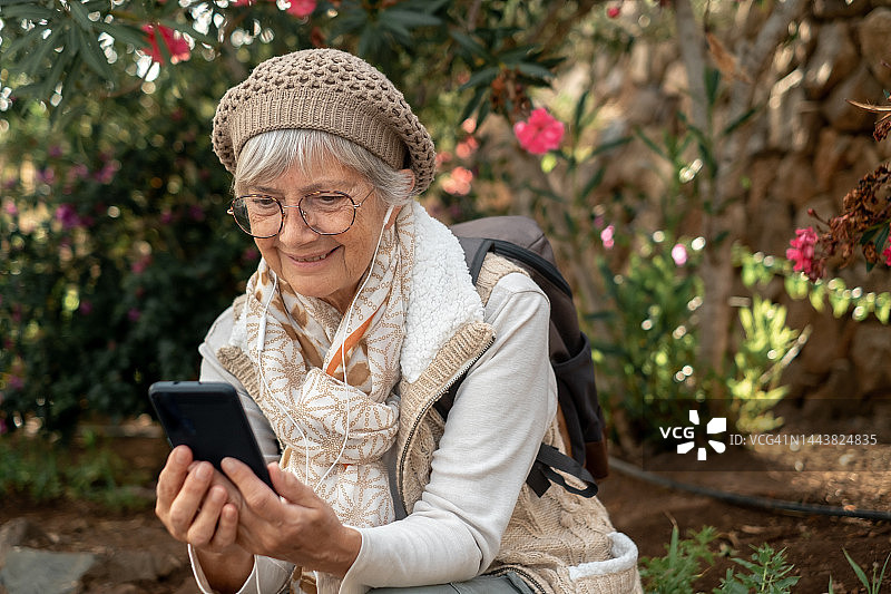 快乐的老年妇女坐在公园里戴着耳机使用手机。美丽的退休女士在户外享受科技和社交图片素材