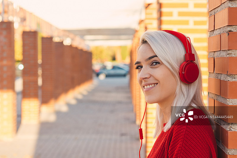 银发女孩戴着红色耳机听音乐。靠在城里的一根柱子上。图片素材