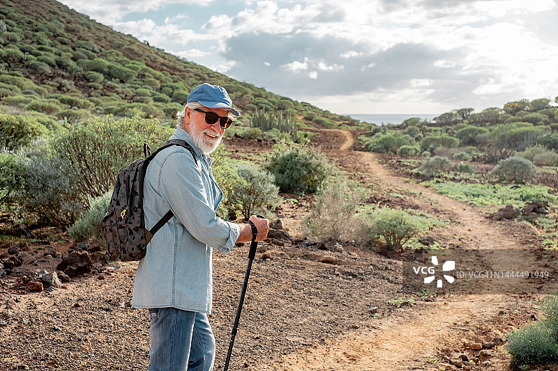 背影的老男子享受徒步在农村沿海。年长的白人男性，戴着帽子，背着背包走在人行道上图片素材