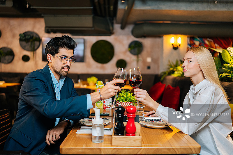 侧视图优雅的年轻情侣在爱碰杯红葡萄酒坐在桌子上在对方在餐厅晚上。图片素材