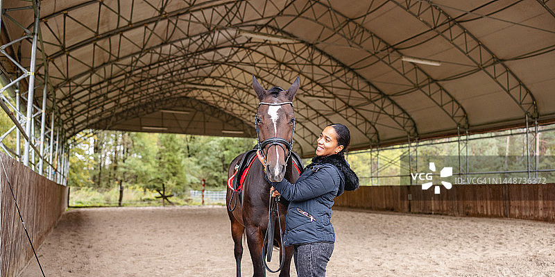 迷人的黑人年轻成年女性爱抚她的棕色马在一个覆盖骑马竞技场图片素材