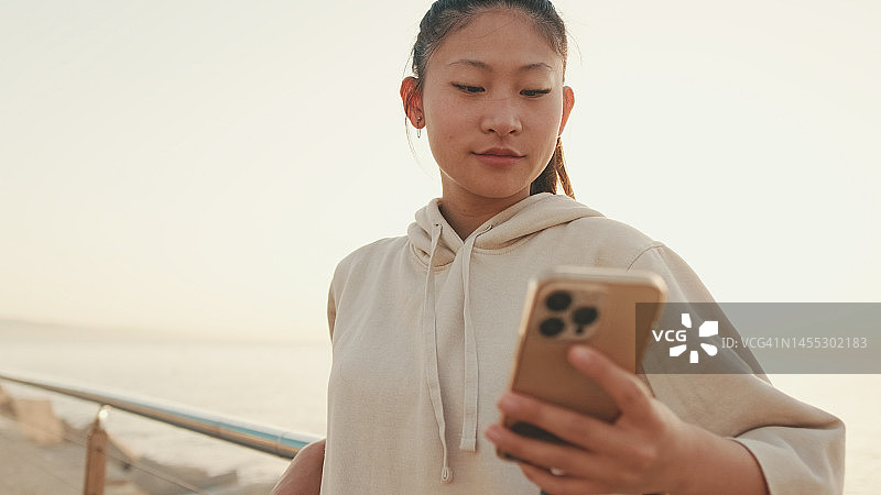 近景是一个穿着运动服的亚洲女孩，早上站在河堤上打电话图片素材