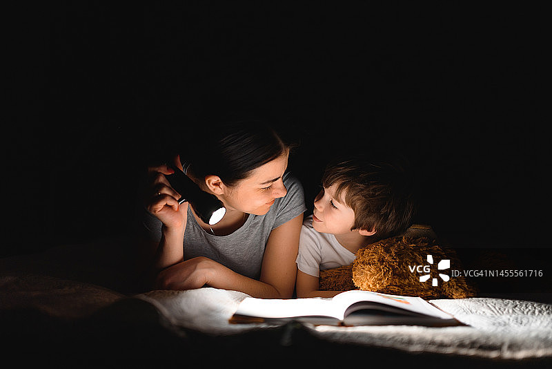 幸福的家庭。是时候讲故事了。快乐可爱的妈妈和儿子在睡觉前享受一本书，并使用手电筒。图片素材