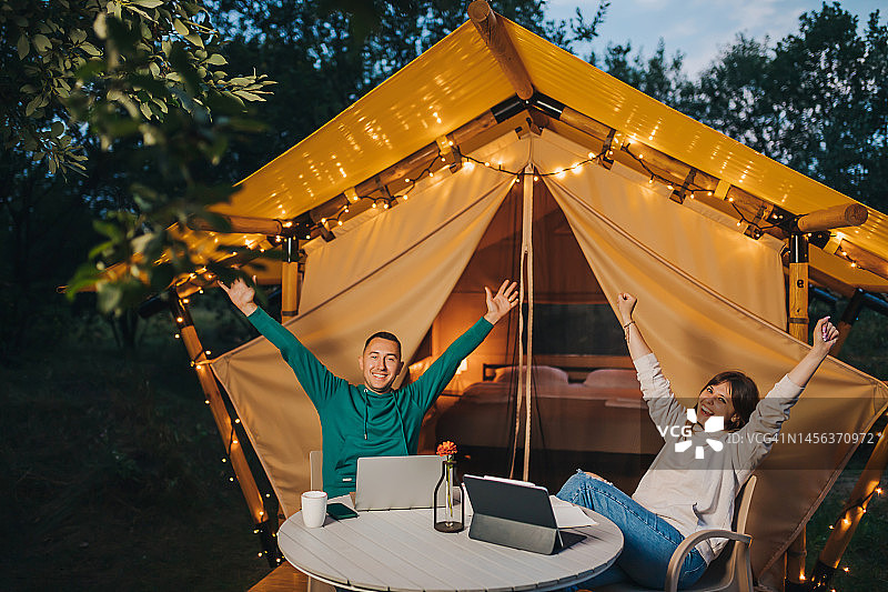 幸福的家庭夫妇自由职业者在一个舒适的露营帐篷在夏夜工作笔记本电脑。户外度假、度假的豪华露营帐篷。生活方式的概念图片素材