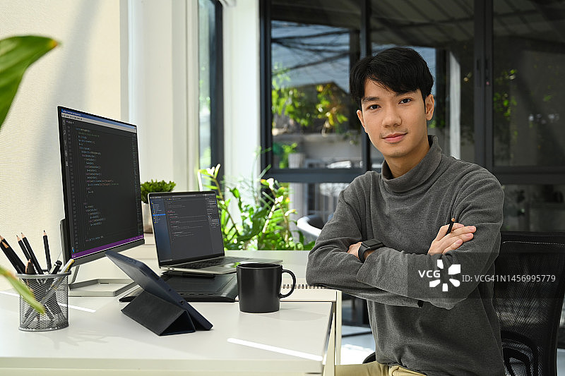 年轻的男性网站开发人员坐在电脑前，屏幕上显示着编码数据，对着镜头微笑。图片素材
