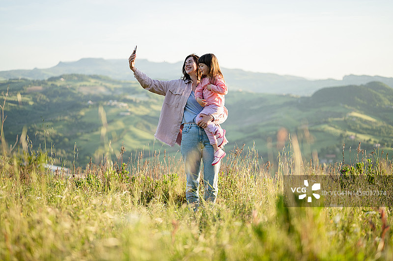 阳光明媚的夏日，在山间草地上自拍。母亲抱着女儿。女人旅行和徒步旅行与蹒跚学步的婴儿。图片素材