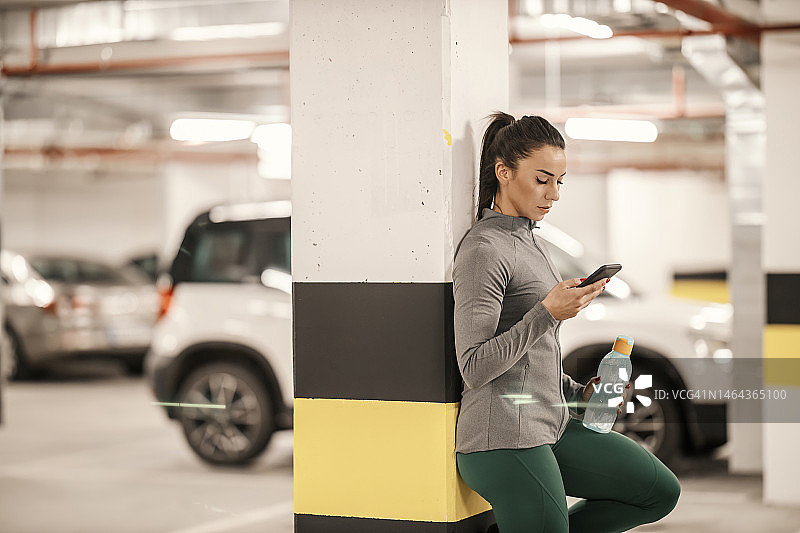 图为，一位健身女运动员在地下车库运动结束后，正在使用手机上的健身应用程序休息。图片素材