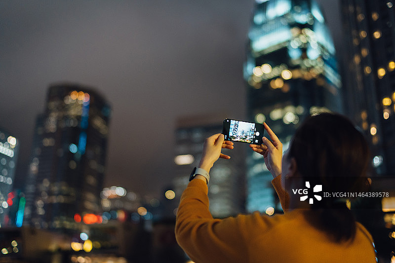 年轻女子用智能手机拍摄城市夜景，对面是市中心明亮多彩的企业摩天大楼图片素材