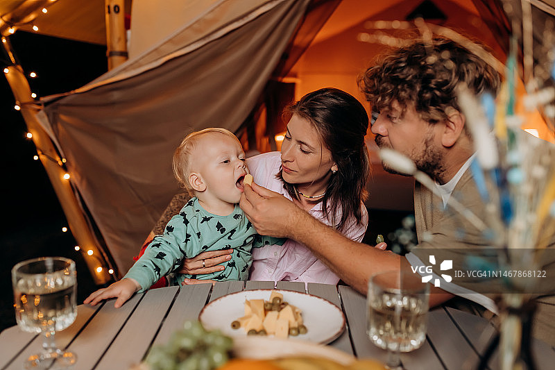 幸福的家庭和可爱的宝宝一起吃晚餐，一起在夏天的晚上在舒适的篝火旁露营。户外休闲休闲的豪华露营帐篷。生活方式的概念图片素材