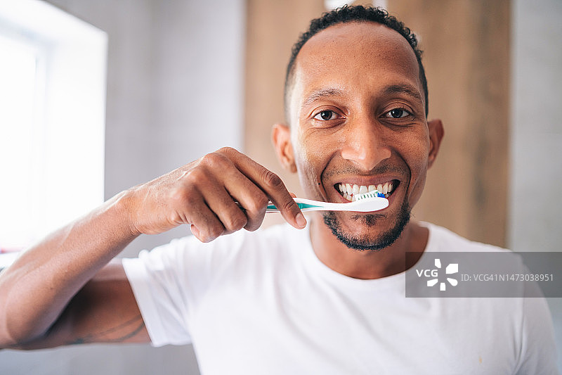 保健，牙齿卫生，人和美丽的概念-微笑的年轻多种族男子用牙刷清洁牙齿，并在家里浴室看镜子图片素材