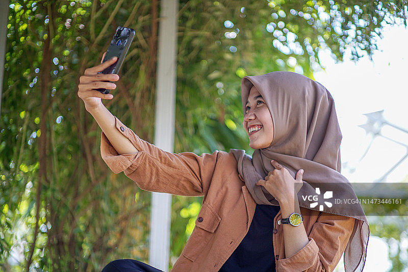 戴着头巾的美丽的亚洲穆斯林女子正在用智能手机自拍图片素材