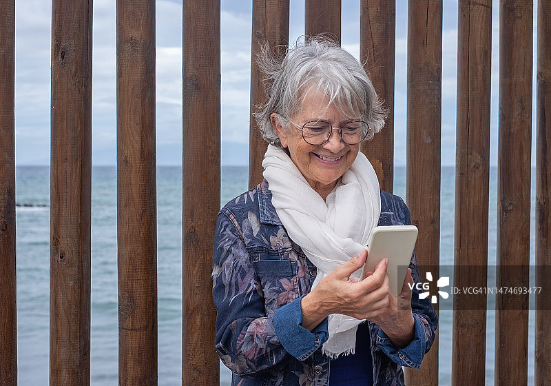 有吸引力的白人老年妇女的肖像与白色围巾坐在户外使用电话。老太太微笑着看智能手机享受自由时间和退休在海上图片素材