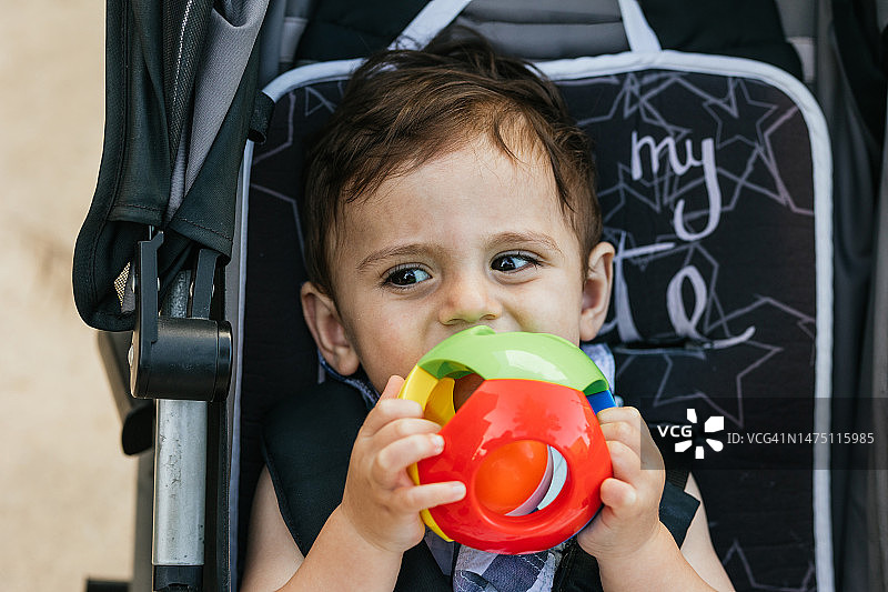 漂亮的小男孩坐在婴儿车上嚼着玩具球。图片素材