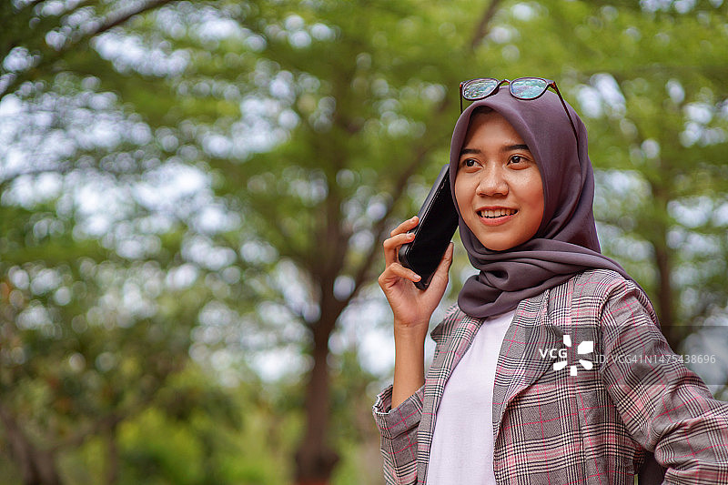 美丽的头巾穆斯林学生打电话给她的朋友在公园用智能手机图片素材
