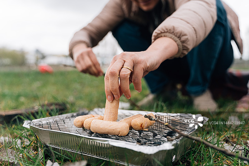 成熟女人的手近距离烹饪香肠在单一使用烤架。在户外野餐图片素材