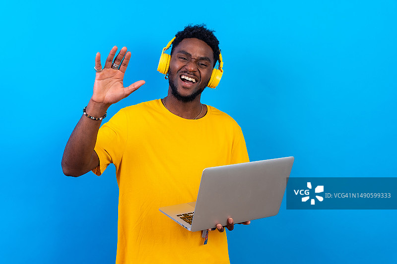 年轻的非洲黑人男子戴着耳机，在蓝色的背景下孤独地跳舞，穿着黄色的t恤，手里拿着电脑，兴奋地享受着舞蹈图片素材