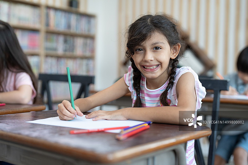 小学美术课上，可爱的小女孩微笑着用彩色铅笔在白纸上画画。儿童教育与学习理念。图片素材