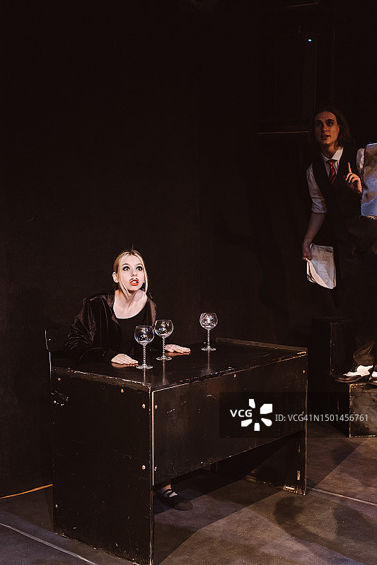 在学生戏剧俱乐部的表演中，一个金发少女坐在桌子旁，手里拿着空杯子，旁边站着一个穿着复古西装的男人图片素材