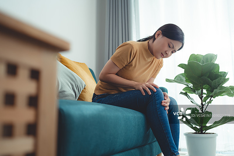 一位亚洲妇女坐在家里客厅的沙发上忍受着膝盖疼痛。图片素材
