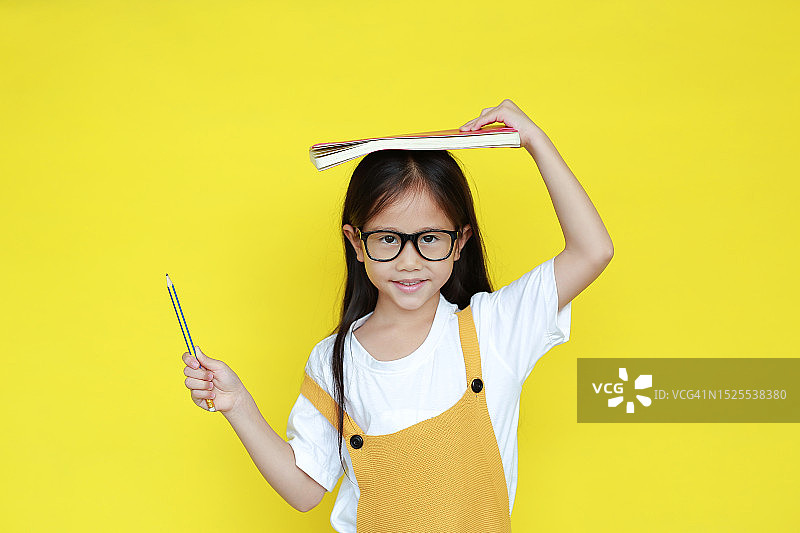 亚洲小女孩戴着眼镜，头上有书，拿着铅笔，微笑着孤立在黄色背景上。肖像快乐的女学生看着相机。学生返校概念图片素材