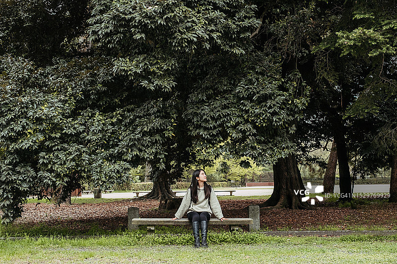 年轻的亚洲女子坐在日本花园的长椅上，靠着树休息。生活方式，旅游，健康理念。图片素材