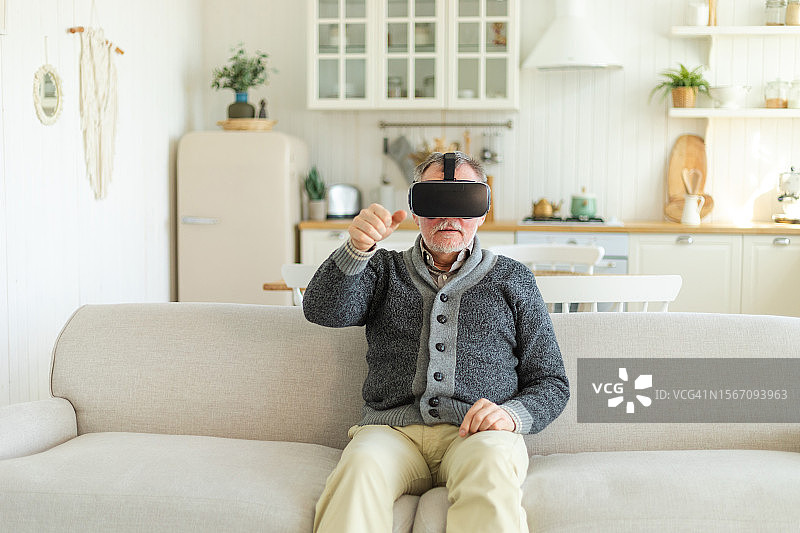 兴奋的成熟老年男子在家中戴着使用虚拟现实的虚拟现实头盔。爷爷在虚拟现实头盔上体验VR时触摸空气。模拟高科技视频游戏。图片素材