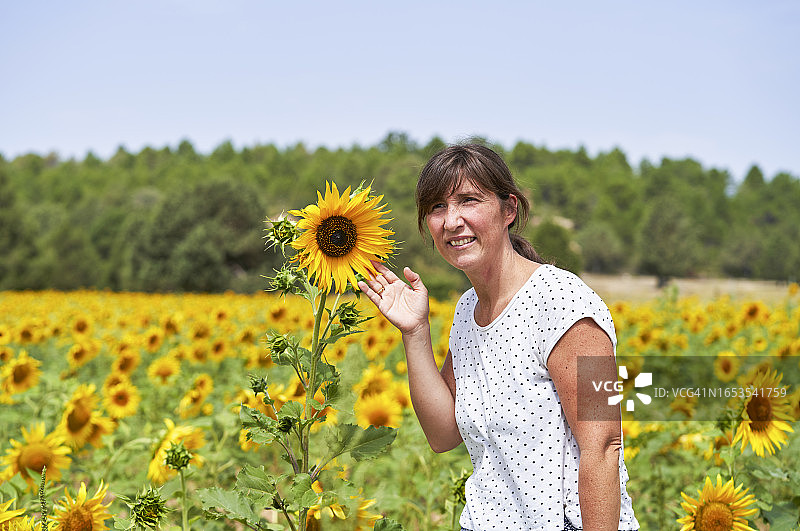 一名身着休闲装的女子站在田野里触摸向日葵，微笑着看着镜头，正面视图图片素材