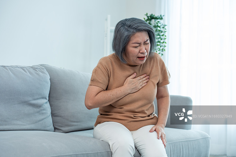 亚洲老年妇女有胸痛的感觉是心脏病发作。有吸引力的老年成熟患者呼吸困难抓着胸口从急性疼痛，而坐在轮椅上的客厅。图片素材
