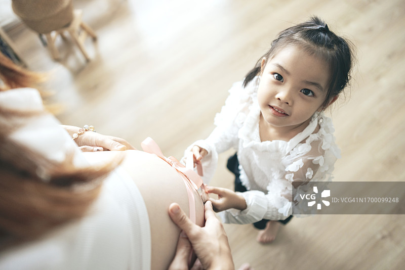 一个年轻的亚洲女孩急切地试图解开她妈妈肚子上的丝带，象征着对新生儿的期待，就像给家人的礼物一样。图片素材