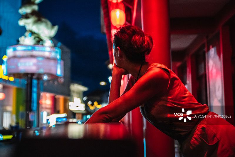 一个孤独的年轻女子倚在栏杆上，身后是红色的霓虹灯，欣赏着马六甲的城市灯光。图片素材