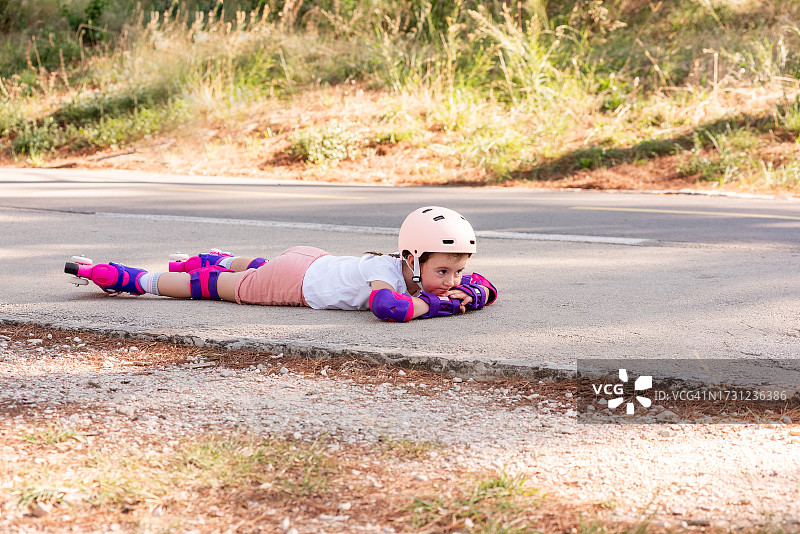 穿着粉色头盔防护服的小女孩躺在柏油路上。孩子害怕滑旱冰图片素材