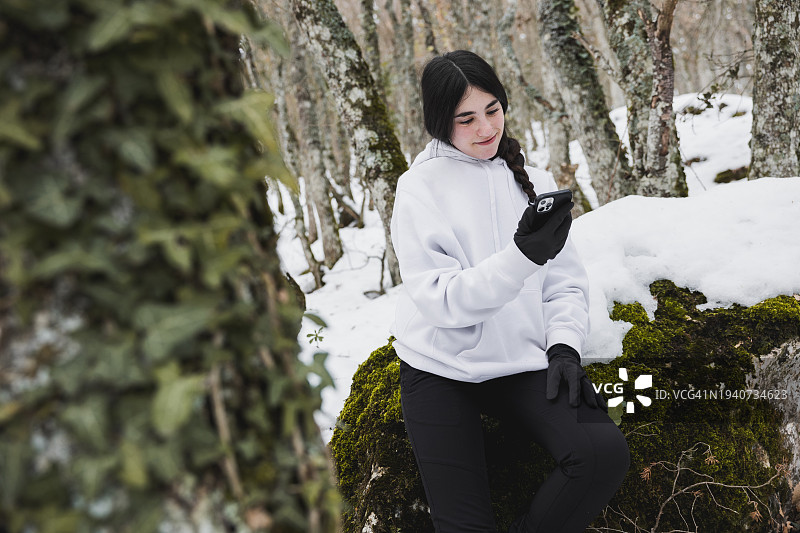 年轻的女孩穿着冬装，在白雪皑皑的森林里拿着手机，在宁静而寒冷的荒野中穿行。图片素材