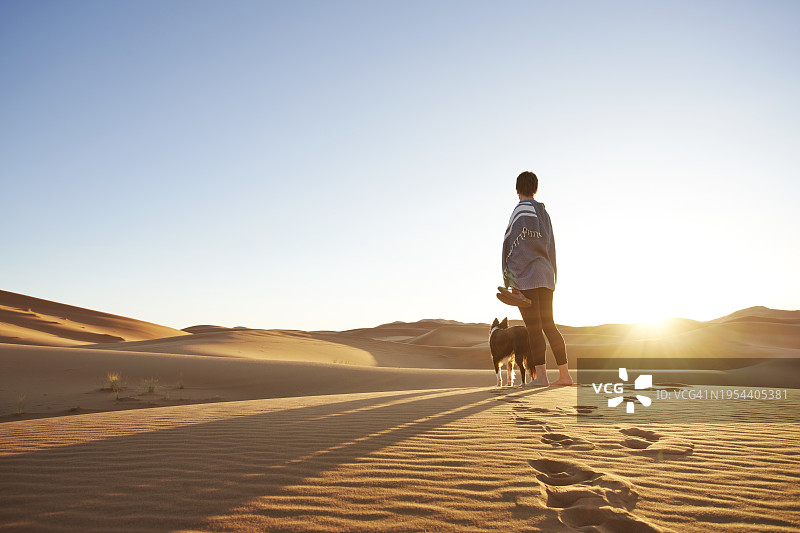 日落时分，孤独的女性和她的狗在沙漠沙丘上散步图片素材