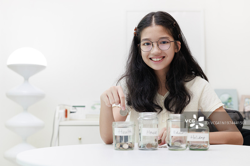 一个年轻的亚洲女孩把硬币放进一个罐子里，象征着她为教育和未来的假期存钱的承诺。图片素材