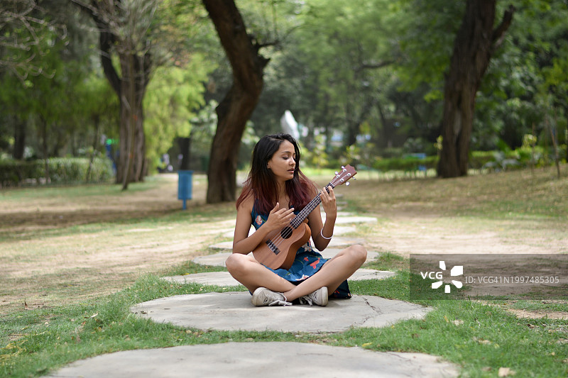 一位年轻的女音乐家，穿着蓝色的花裙，坐在花园里弹着尤克里里琴。图片素材
