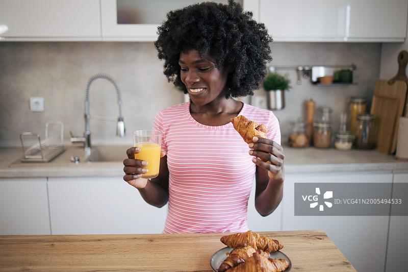 微笑的非裔美国妇女站在餐桌前，早上喝着一杯果汁，吃着一个羊角面包。图片素材