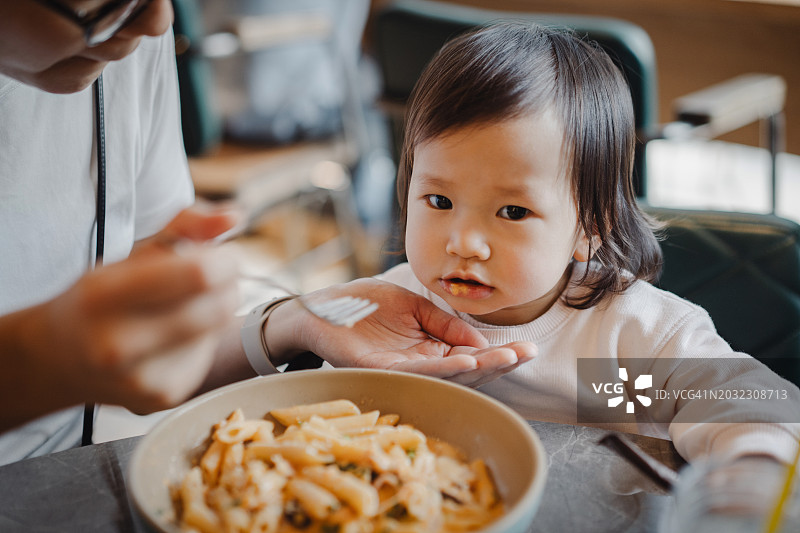一个可爱的年轻亚洲母亲在餐馆里和她蹒跚学步的女儿分享意大利面，在一个舒适的下午愉快地一起吃饭。全家外出就餐的生活方式。亲情之爱，关怀与纽带的理念图片素材