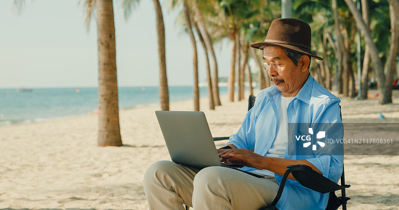 一位亚洲老人坐在沙滩椅上欣赏海景。一个快乐的成年老人享受退休后的旅行生活方式。图片素材