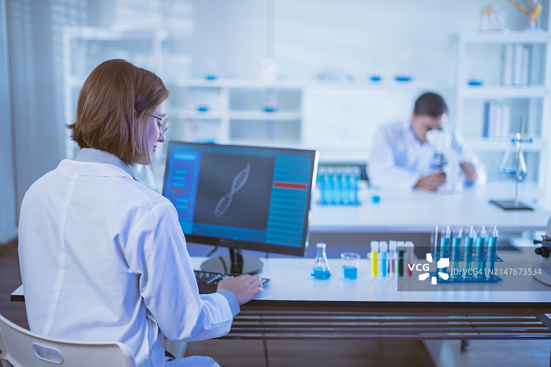 科学家在实验室分析电脑屏幕上的DNA数据，同事在后台工作。研究和生物技术概念。图片素材