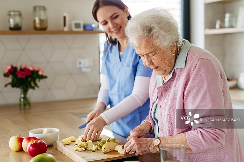 家庭护理人员和老妇人切苹果，做健康快餐。老年妇女做家务，护士帮忙。图片素材