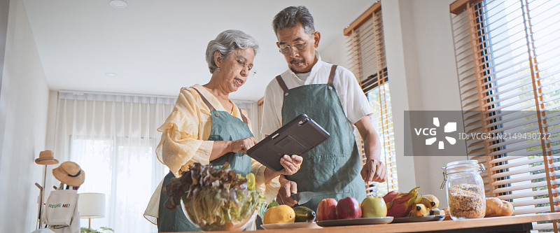 亚洲老年夫妇在家厨房一起做饭，用数码平板电脑制作沙拉菜，用水果、蔬菜烹饪有机食品。科技让人生活方式，健康幸福图片素材