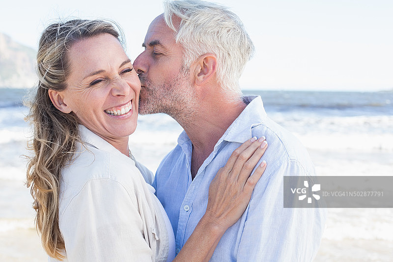 一名男子在海滩上亲吻微笑着的伴侣的脸颊图片素材
