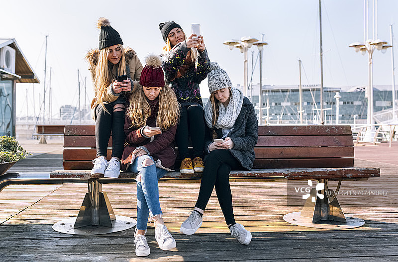 四个年轻女人坐在长椅上玩手机图片素材