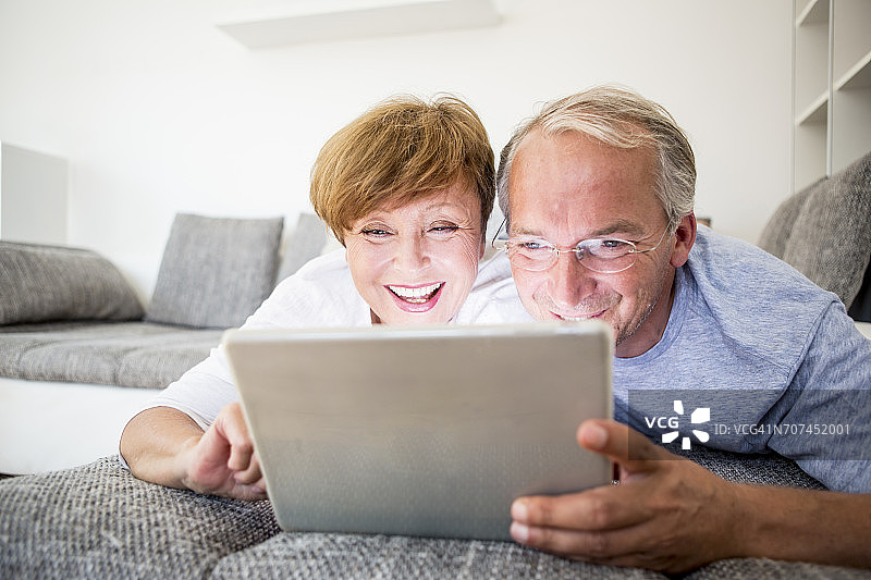 一对快乐的老年夫妇躺在家里的沙发上使用数字平板电脑图片素材