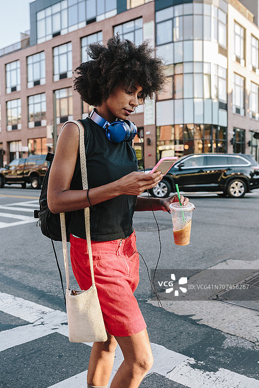 一名年轻女子带着耳机和智能手机穿过布鲁克林大街，随身携带酒水图片素材