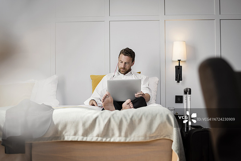 一个商人坐在床上靠墙用笔记本电脑在酒店房间图片素材