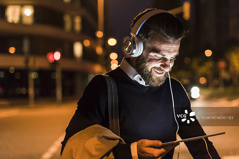 微笑的年轻人，带着平板电脑和耳机在城市的街道上图片素材