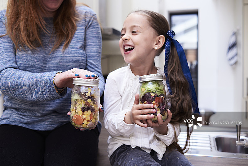 笑着的小女孩拿着一罐混合沙拉坐在她妈妈旁边图片素材